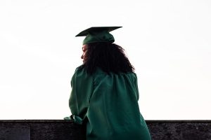 A woman leaning on a wall against a white background dressed in graduation regalia