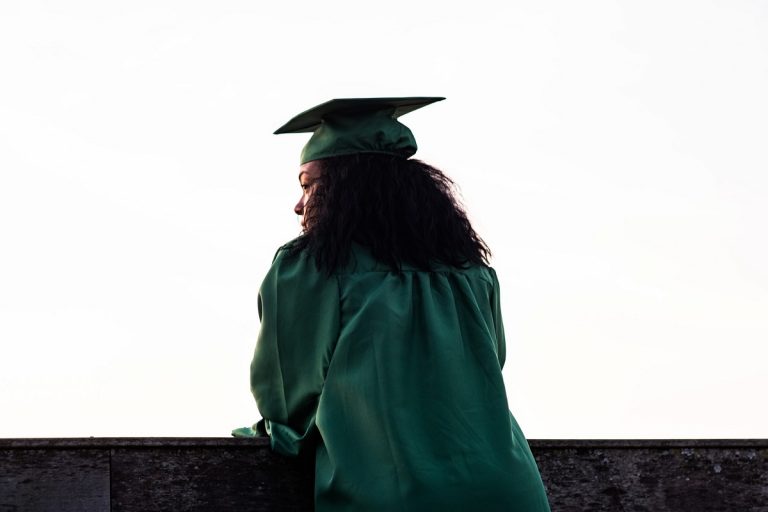 A woman leaning on a wall against a white background dressed in graduation regalia