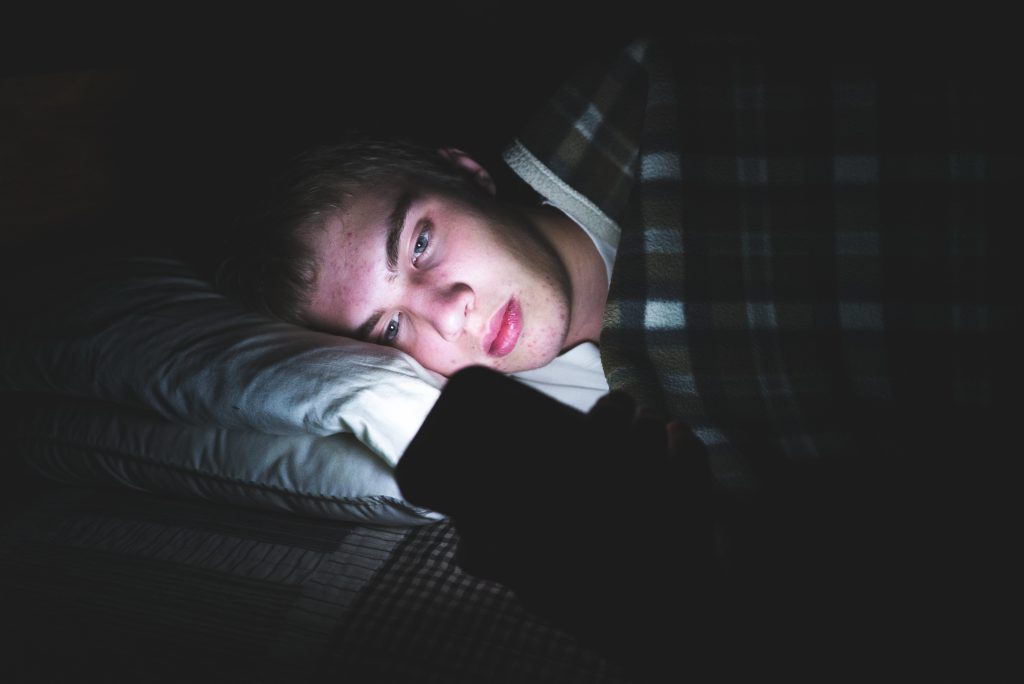 Depressed teenager browsing the internet on his mobile phone as he is lying on his bed in the dark.