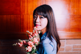 Portrait of poet Jane Wong, who is holding a bouquet of flowers and standing in front of a wood paneled wall.